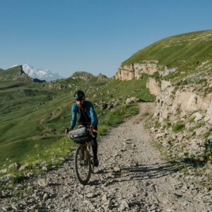 Un ciclista con abbigliamento tecnico sportivo e bici da gravel equipaggiata per il bikepacking pedala su una strada sterrata di montagna in primavera, con prati verdi e una vetta innevata sullo sfondo.