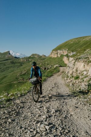 Un ciclista con abbigliamento tecnico sportivo e bici da gravel equipaggiata per il bikepacking pedala su una strada sterrata di montagna in primavera, con prati verdi e una vetta innevata sullo sfondo.
